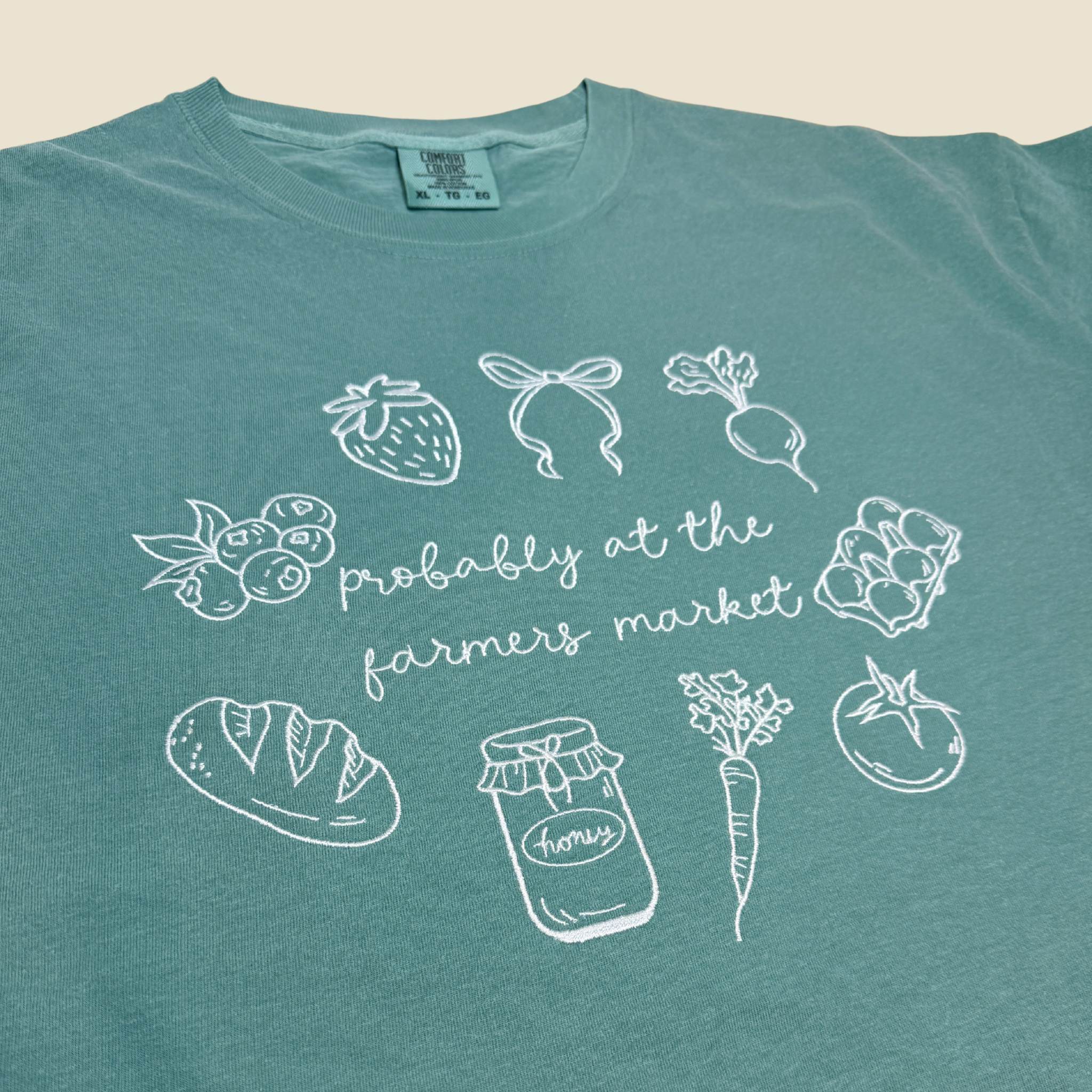 Light green t-shirt with white illustrations and text 'probably at the farmers market' on a beige background