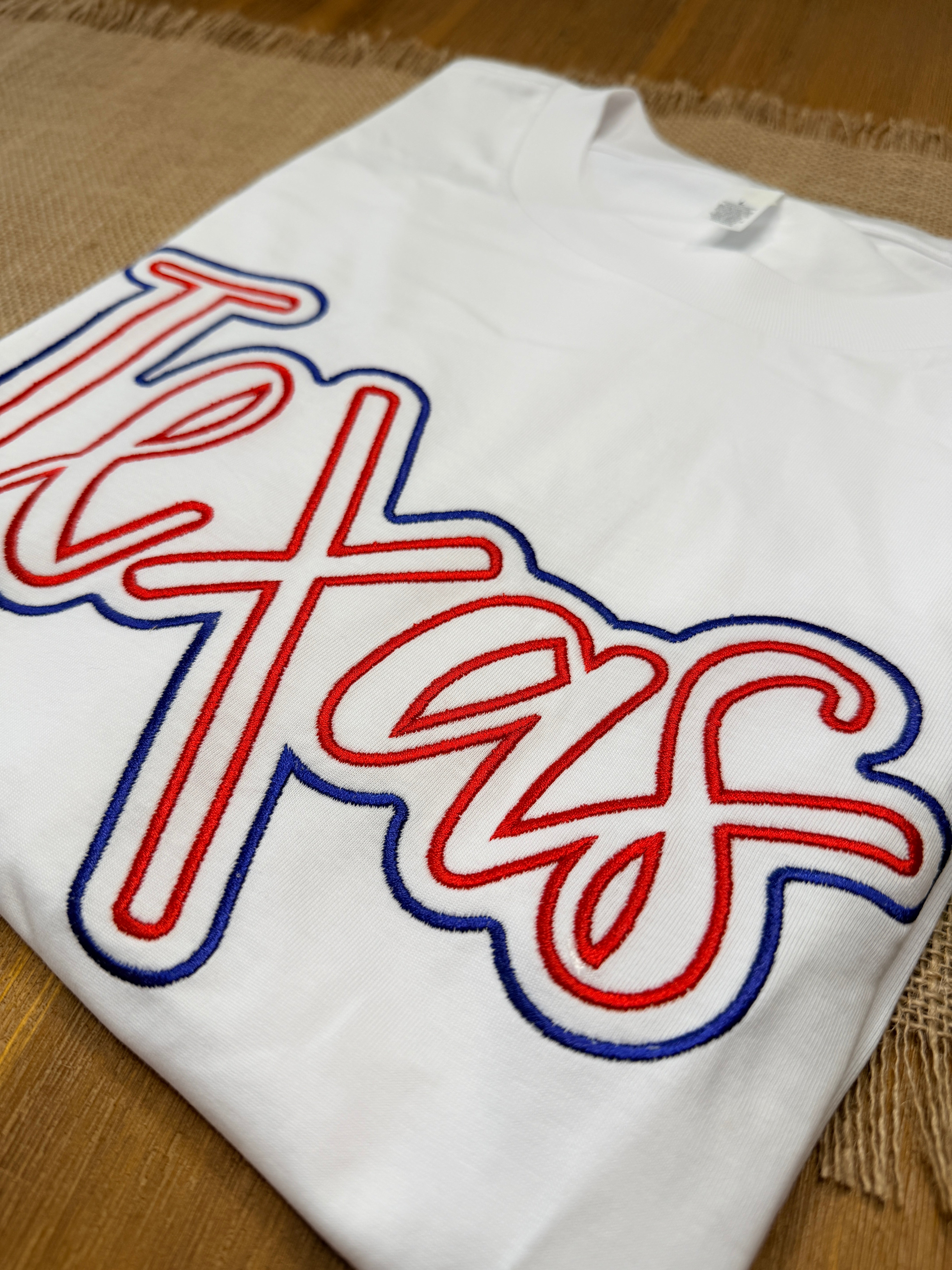 White t-shirt with red and blue 'Texans' logo on a wooden surface