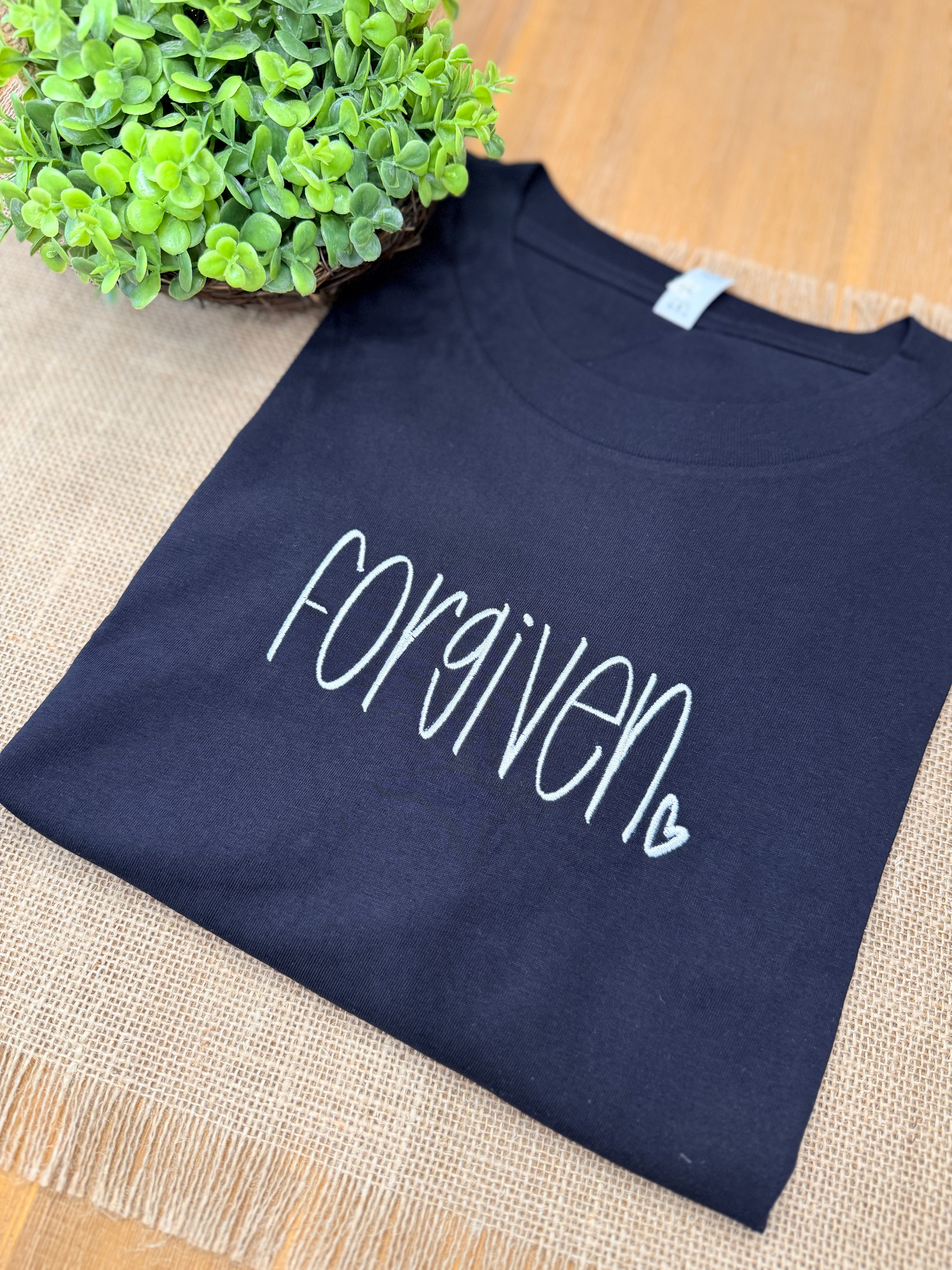 black t-shirt with 'Forgiven' text on a textured surface with a plant in the corner.