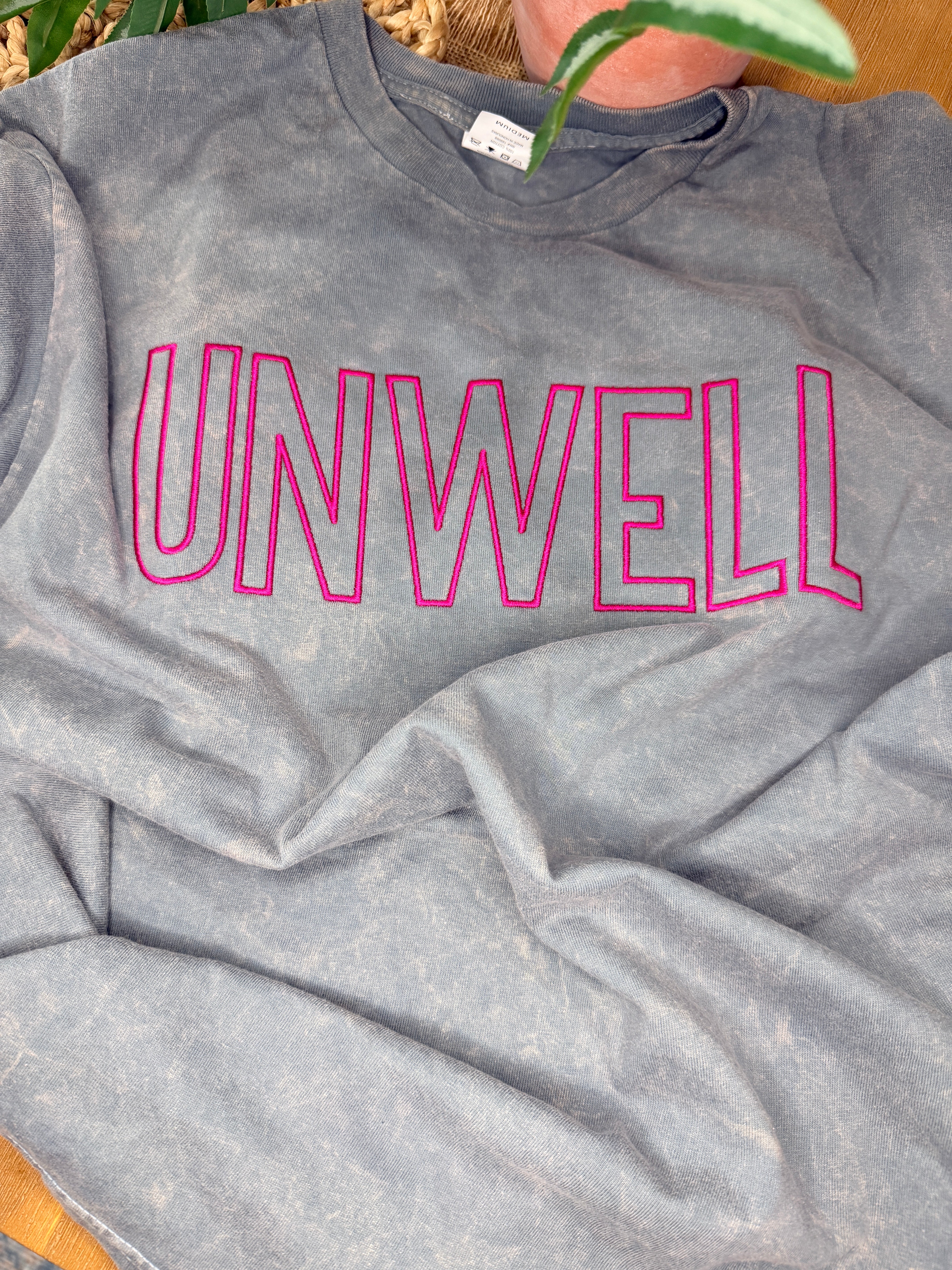 Gray acid-washed Cotton Collective tour tee with bold pink “UNWELL” embroidery across the chest. Relaxed fit with soft vintage wash and minimalist statement design.