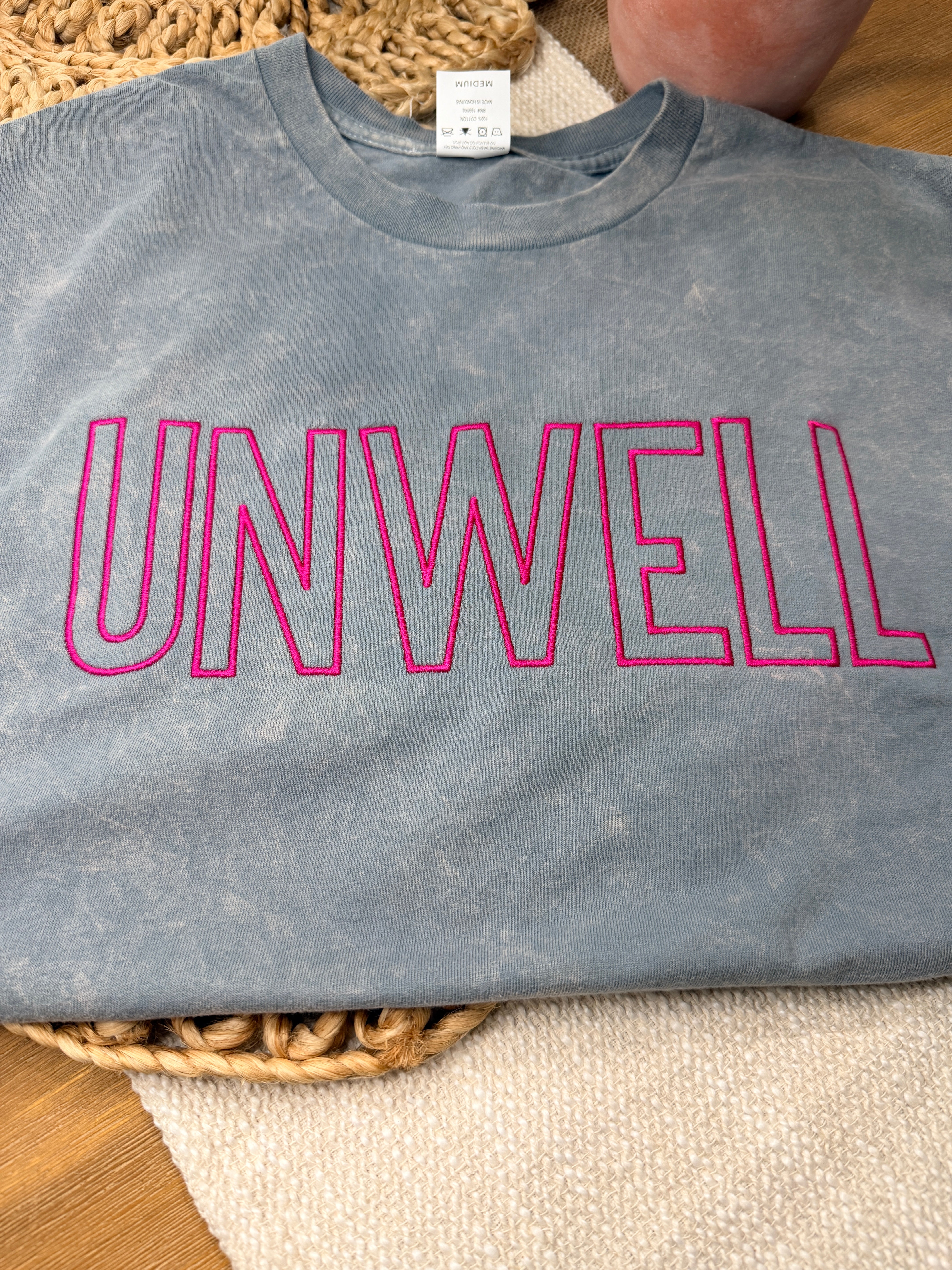 Gray acid-washed Cotton Collective tour tee with bold pink “UNWELL” embroidery across the chest. Relaxed fit with soft vintage wash and minimalist statement design.