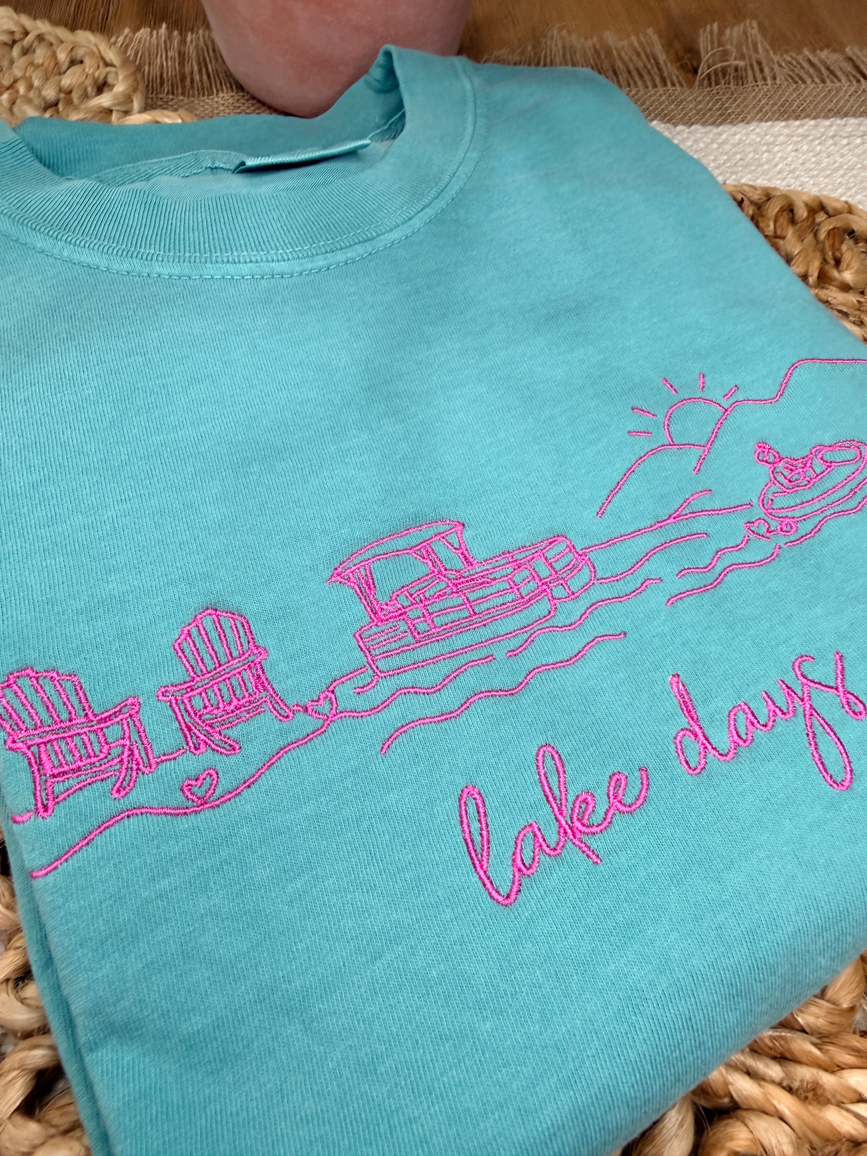 Seafoam Comfort Colors tee with pink line embroidery of lake scene including chairs, dock, boat, and sun, plus “lake days” script. Relaxed fit with soft garment-dyed finish.