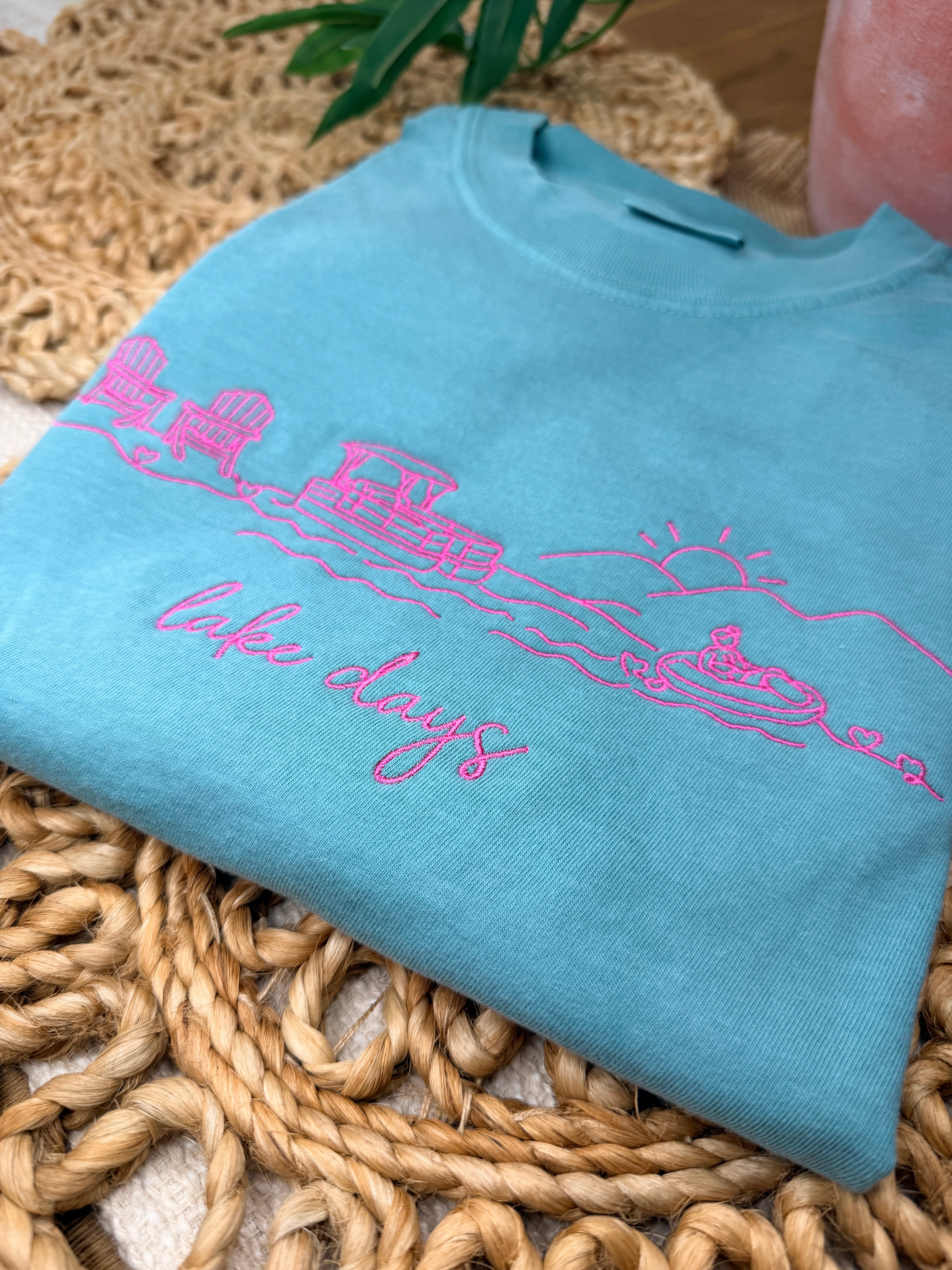 Seafoam Comfort Colors tee with pink line embroidery of lake scene including chairs, dock, boat, and sun, plus “lake days” script. Relaxed fit with soft garment-dyed finish.