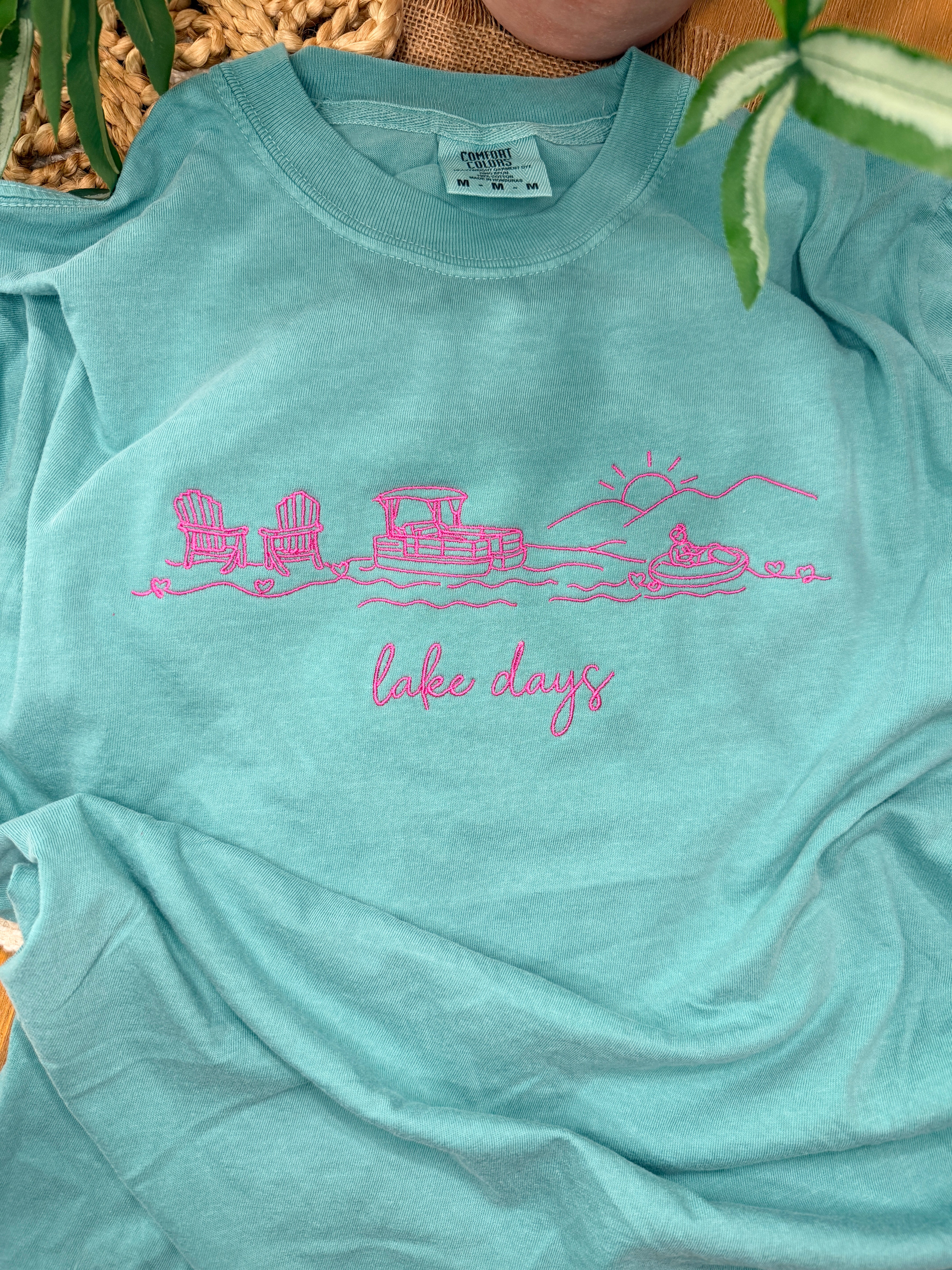 Seafoam Comfort Colors tee with pink line embroidery of lake scene including chairs, dock, boat, and sun, plus “lake days” script. Relaxed fit with soft garment-dyed finish.