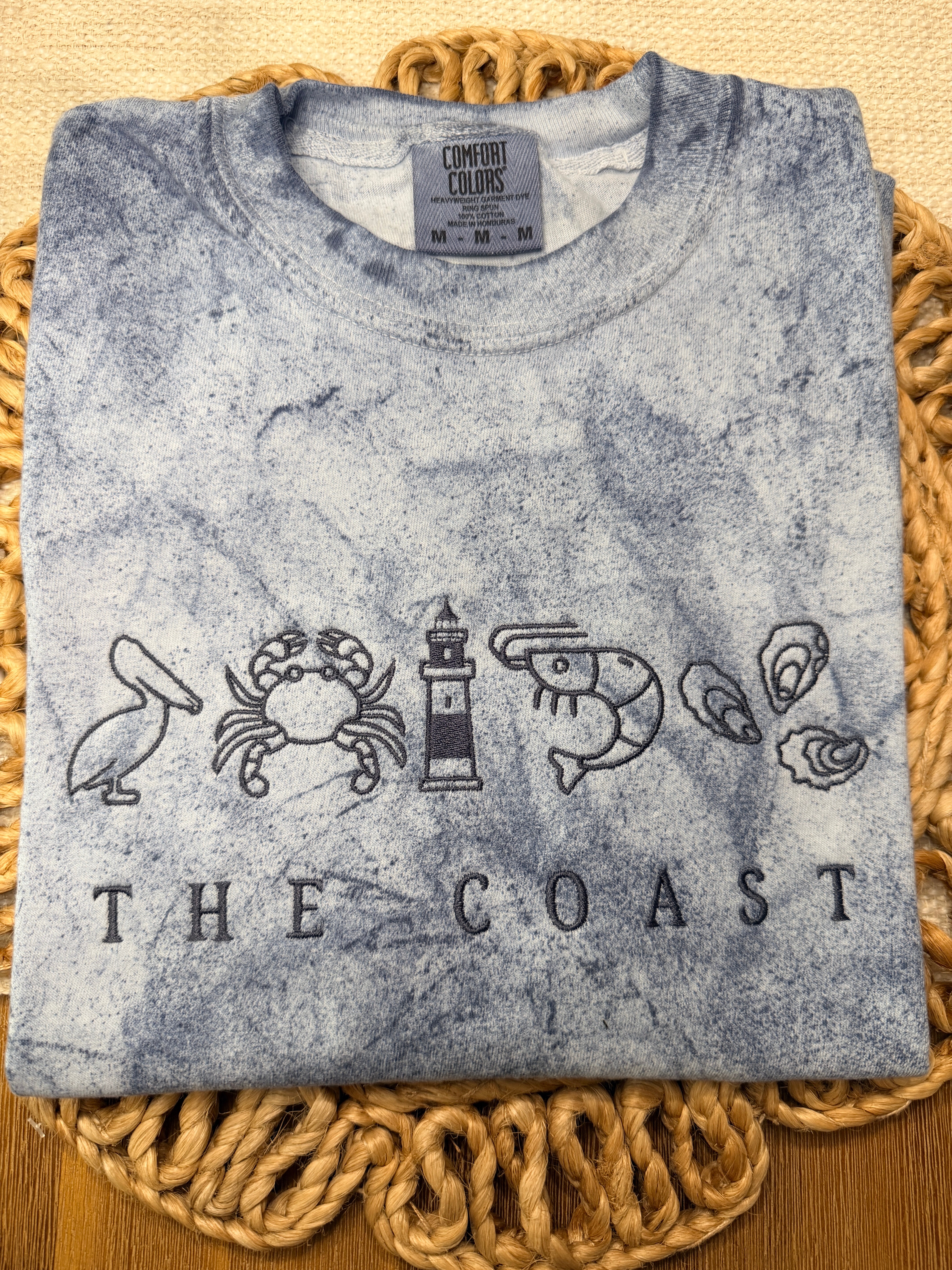 Blue-gray Comfort Colors Color Blast tee with coastal embroidery featuring pelican, crab, lighthouse, shrimp, and oysters above “THE COAST” text. Relaxed fit with unique washed pattern.