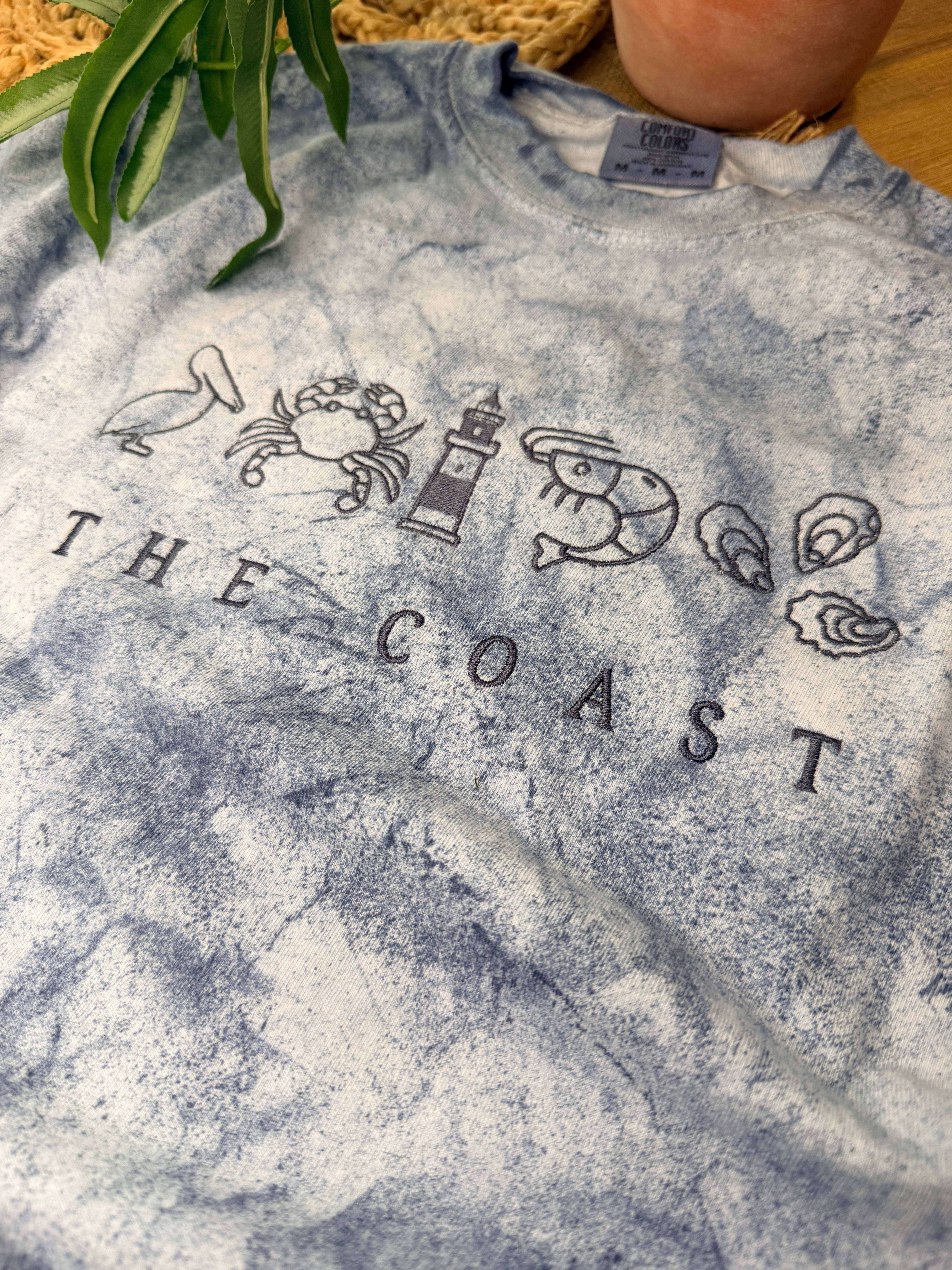 Blue-gray Comfort Colors Color Blast tee with coastal embroidery featuring pelican, crab, lighthouse, shrimp, and oysters above “THE COAST” text. Relaxed fit with unique washed pattern.