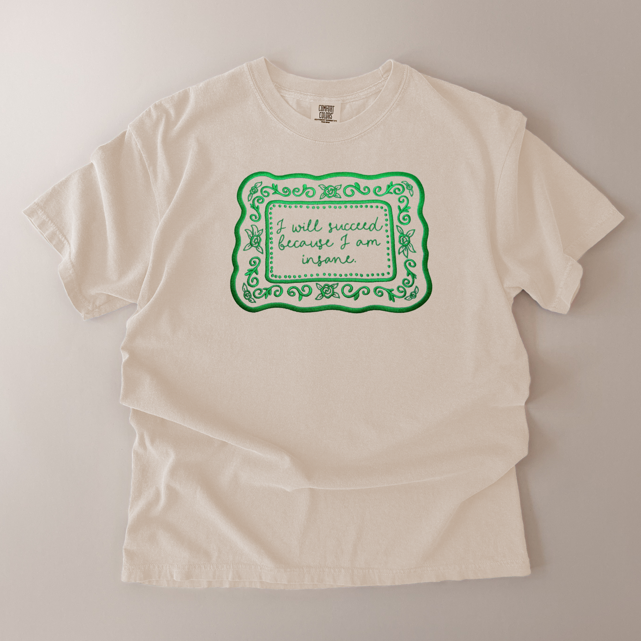 Beige t-shirt with green text and decorative elements on a light gray background