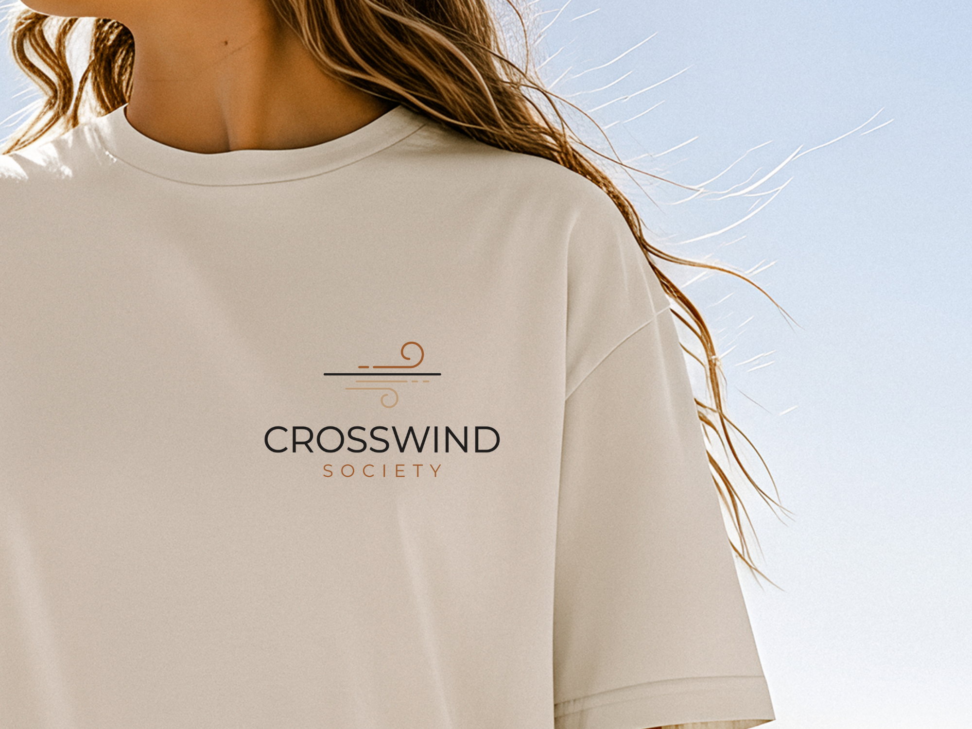 Person wearing a beige sweatshirt with 'Crosswind Society' logo against a clear blue sky.
