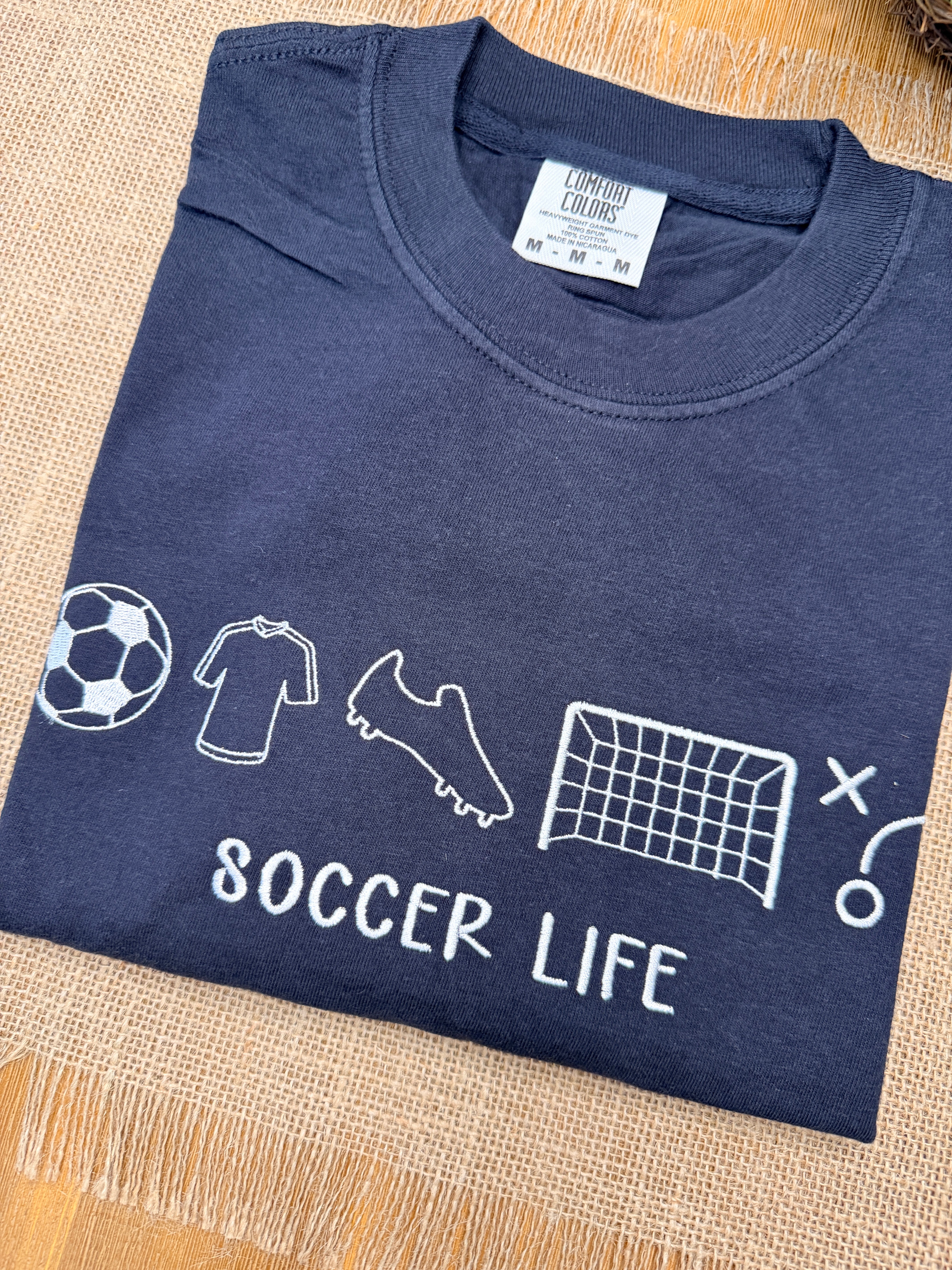 Folded navy blue t-shirt with white graphics and 'Soccer Life' text on a beige surface