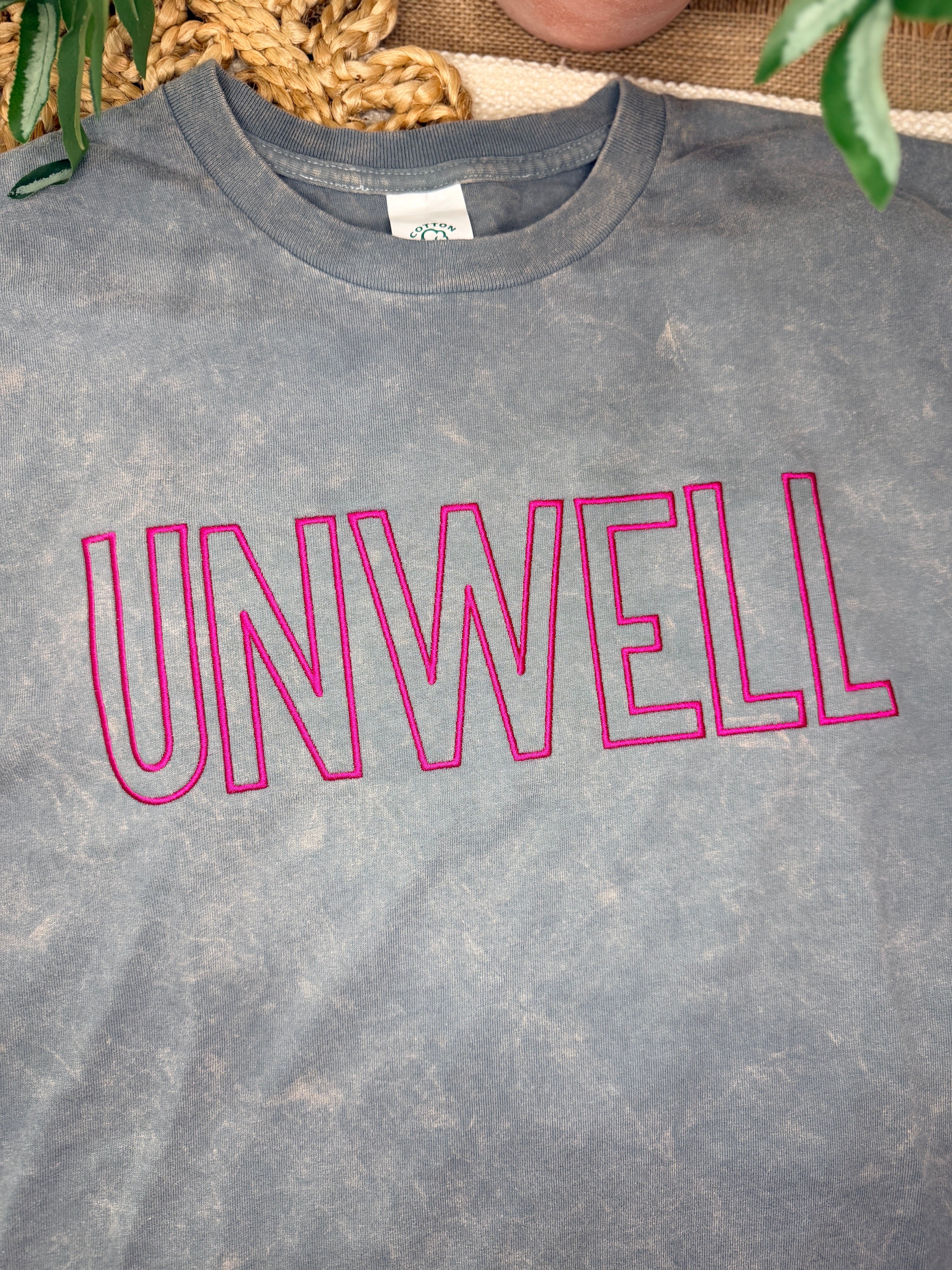 Gray acid-washed Cotton Collective tour tee with bold pink “UNWELL” embroidery across the chest. Relaxed fit with soft vintage wash and minimalist statement design.