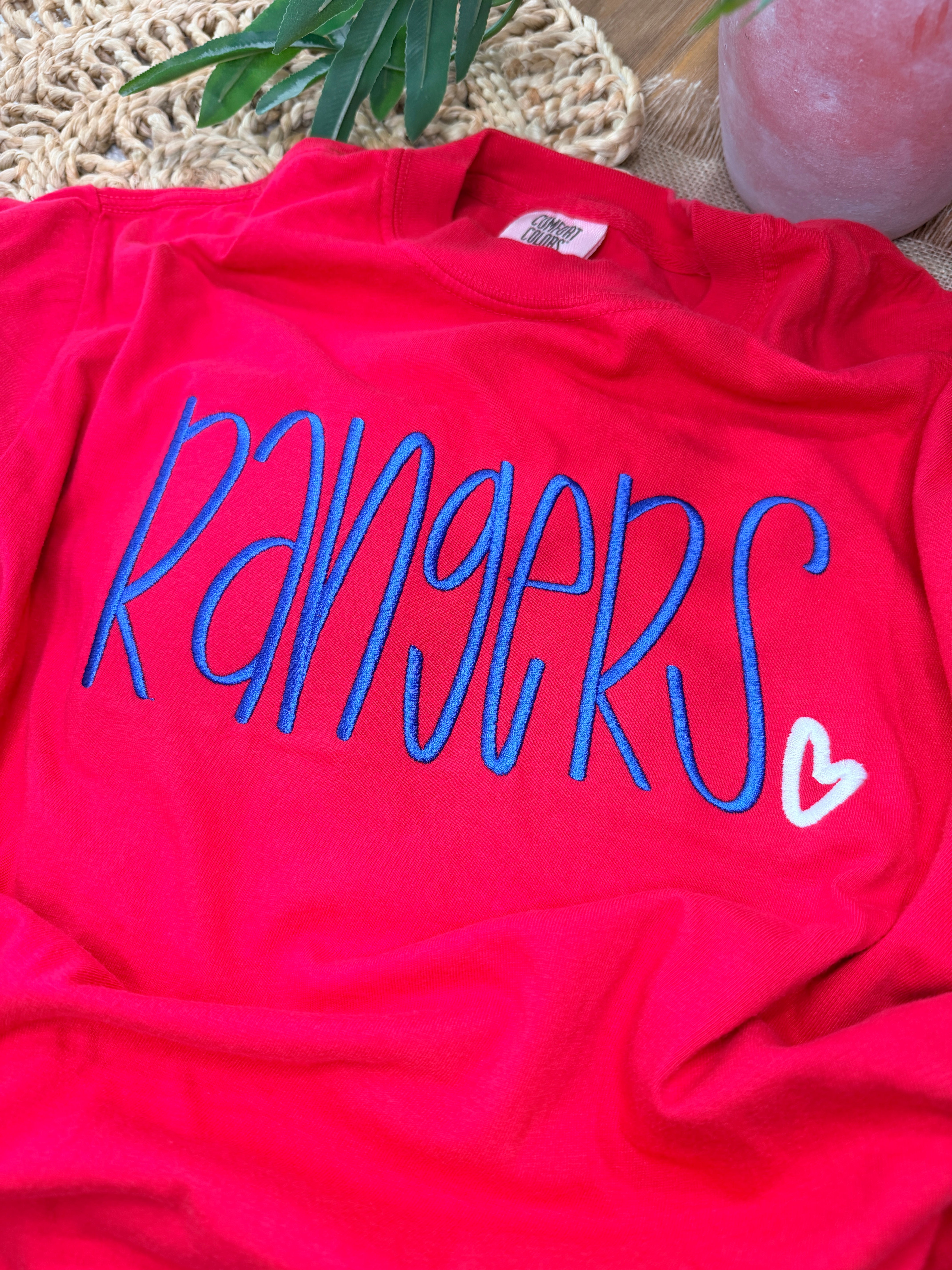 Red Comfort Colors tee with large blue “Rangers” script embroidery and small white heart detail. Soft garment-dyed cotton with relaxed unisex fit and bold front design.