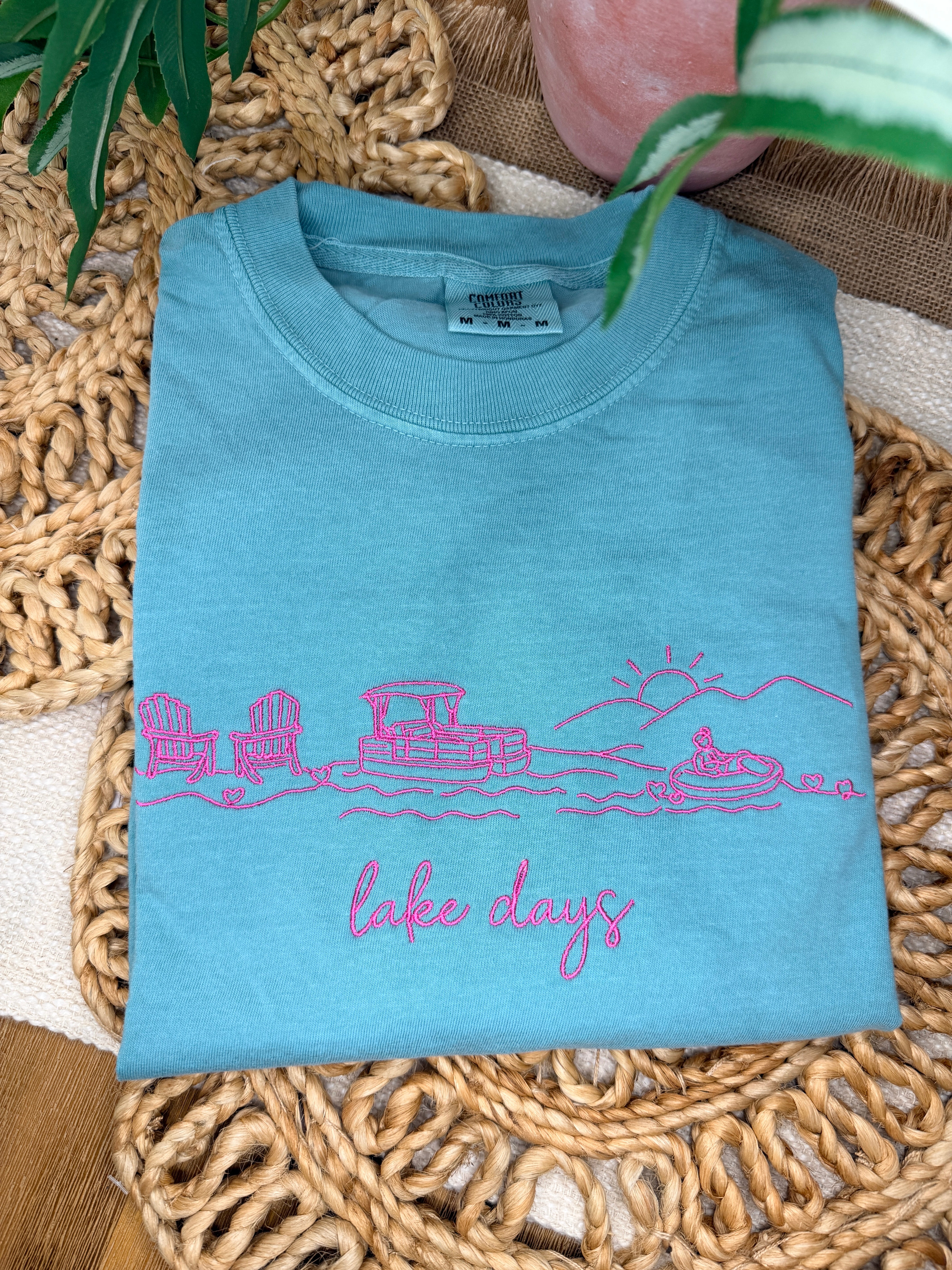 Seafoam Comfort Colors tee with pink line embroidery of lake scene including chairs, dock, boat, and sun, plus “lake days” script. Relaxed fit with soft garment-dyed finish.