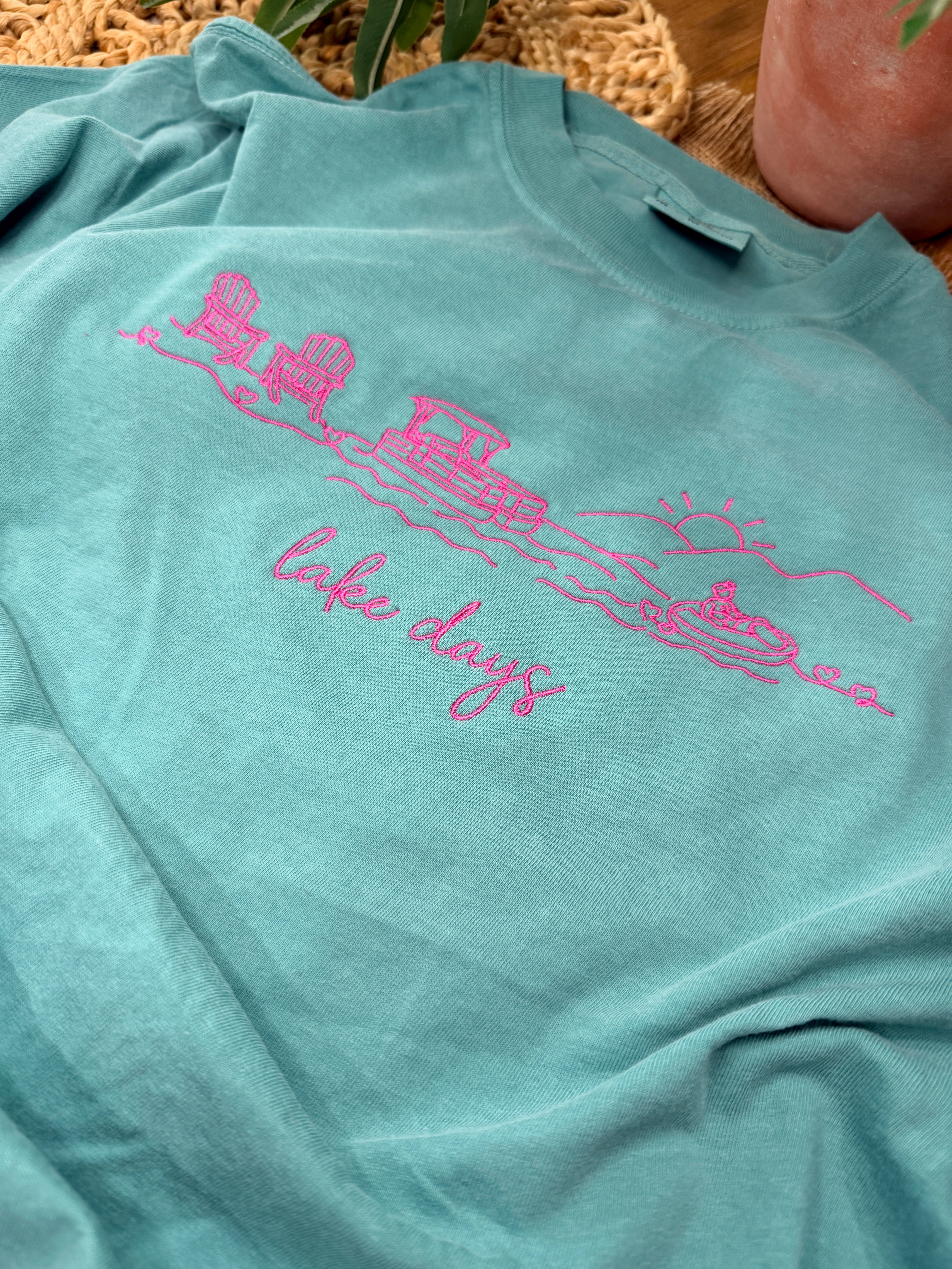 Seafoam Comfort Colors tee with pink line embroidery of lake scene including chairs, dock, boat, and sun, plus “lake days” script. Relaxed fit with soft garment-dyed finish.