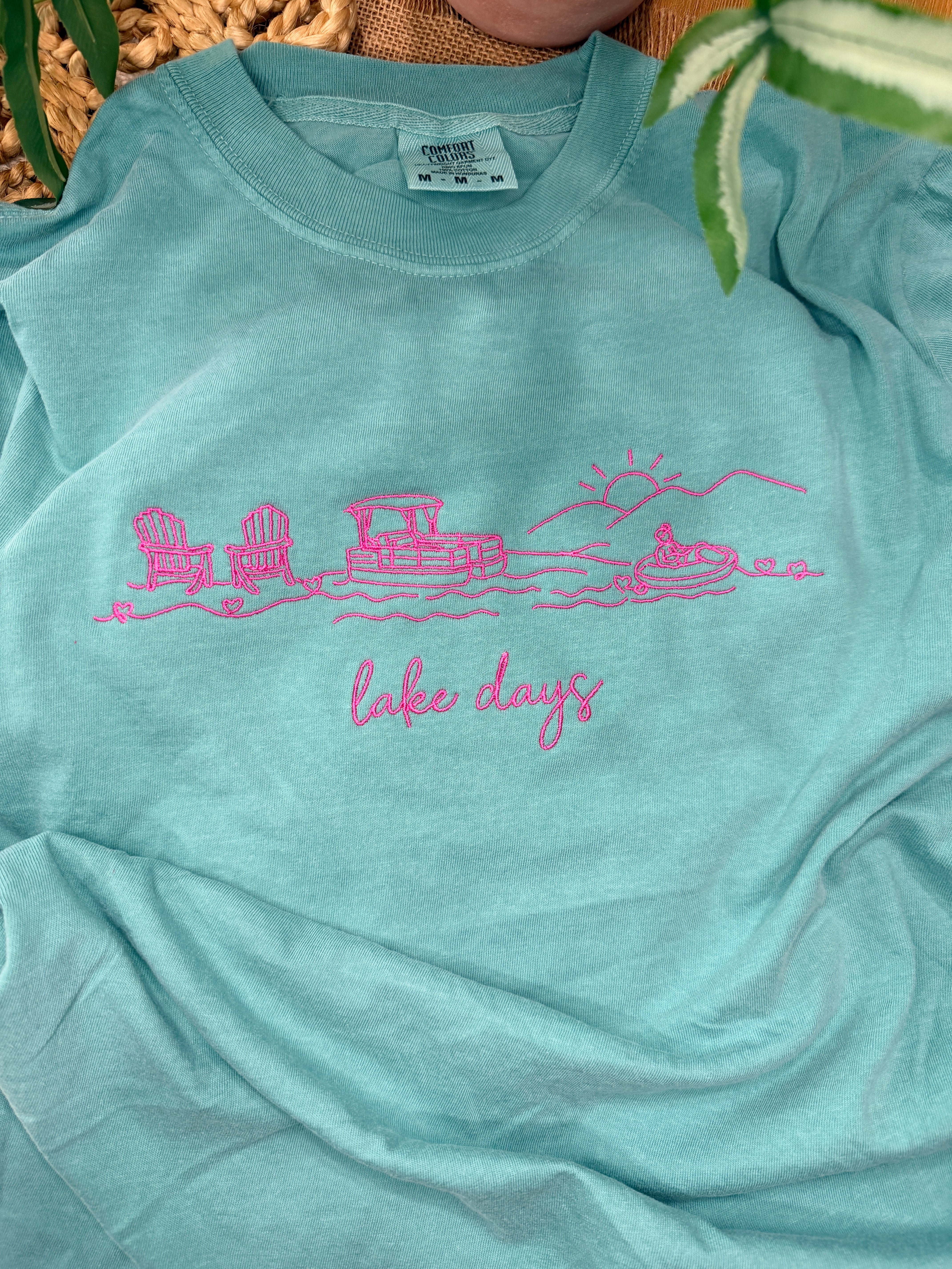 Seafoam Comfort Colors tee with pink line embroidery of lake scene including chairs, dock, boat, and sun, plus “lake days” script. Relaxed fit with soft garment-dyed finish.