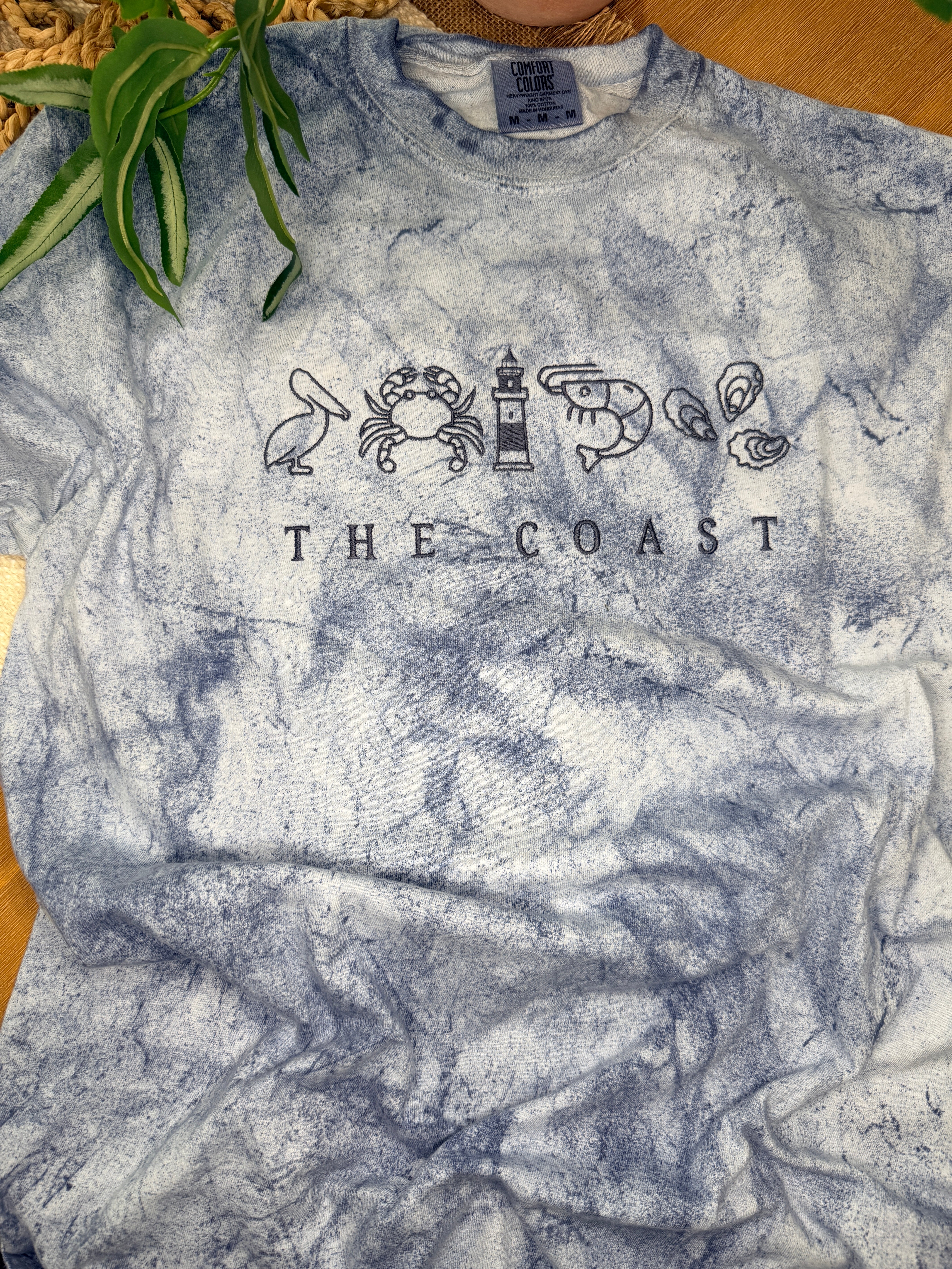 Blue-gray Comfort Colors Color Blast tee with coastal embroidery featuring pelican, crab, lighthouse, shrimp, and oysters above “THE COAST” text. Relaxed fit with unique washed pattern.